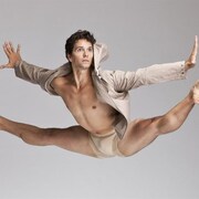 Le danseur Guillaume Côté est posé alors qu'il fait le grand écart dans les airs. On comprend qu'il a jumelé un saut et la figure du grand écart. l'artiste est vêtu d'une culotte et manteau court beige.