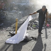 Un homme lève le drap recouvrant la victime d'une fusillade dans une rue jonchée de déchets.