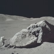 Une photo en noir et blanc d'un astronaute sur la Lune en 1972. Il semble tout petit et est debout faisant face à un très gros rocher.