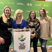 Sami Jo Small, Heather Morrison, Vicki Keith et Heather Moyse posent en souriant pour une photo entre un grand mur vert et un podium décoré du logo des Jeux du Canada 2023.