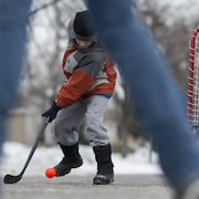 Des enfants jouent au hockey dans la rue en hiver. Un garçon d'âge primaire tente de contrôler une balle rouge avec un bâton de hockey. Une fillette est devant un but posé dans la rue.
