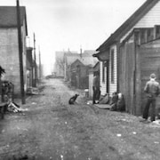 De petites maisons de bois le long d'une ruelle de terre où est assis un chien. Un homme rentre chez lui alors qu'un autre marche près des maisons du côté droit de la rue.