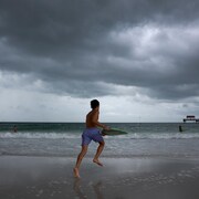 Un jeune homme en maillot court sur une plage, avec une planche de surf dans les mains, alors qu'il fait sombre.