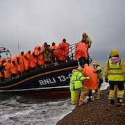 Un bateau accoste sur une plage. A son bord, une vingtaine d'hommes vêtus de cirés orange. Sur la plage de galets, des hommes portant une veste jaune sur laquelle est écrit Shore Crew. 
