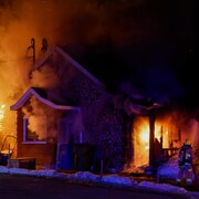 Une maison est en proie aux flammes. On voit un pompier qui tente d'éteindre le brasier devant un nuage de fumée qui sort de la maison.