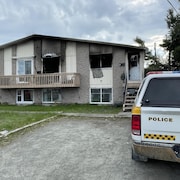 Une voiture de la Sûreté du Québec est stationnée devant une maison endommagée par un incendie. Les fenêtres et la porte sont détruites et des traces de fumée apparaissent sur le devant de la maison.