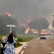 Une femme debout près d'une moto prend une photo d'un incendie à côté d'une route sur laquelle circule une voiture beige. 