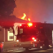 Un camion de pompier devant une maison où on voit des flammes sortir du toit.