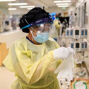 Une travailleuse de la santé porte une visière de protection médicale, des lunettes, et un masque. Elle regarde un gant. 