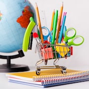 Un panier d'épicerie rempli de crayons, d'effaces et de ciseaux est posé sur des cahiers de notes devant un globe terrestre.