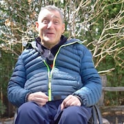 Un homme en fauteuil roulant fait face à la caméra dans un sentier nature devant des arbres. 