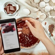 Une personne saupoudre de cacao un gâteau au chocolat en lisant les indications de la recette sur son cellulaire. 