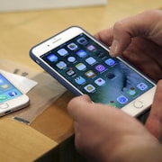 Plan rapproché des mains d'une personne manipulant un iPhone 7 Plus, alors qu'un iPhone 6 est posé sur une table en arrière-plan.
