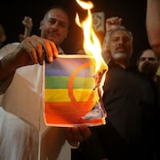 Un homme dans une foule tient une feuille en train de brûler avec le drapeau LGBTQ+ avec un signe rouge d'interdiction dessus.