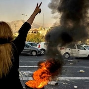 Une femme fait un signe de la main devant une voiture en flammes.
