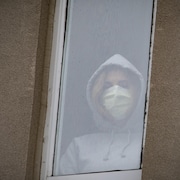 Une jeune femme regarde à travers la fenêtre d'un hôpital, portant un masque.