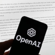 Une main tient un téléphone intelligent affichant le logo d'OpenAI, devant un écran d'ordinateur affichant l'outil ChatGPT. 