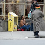 Un homme est assis sur le trottoir, torse nu, tandis que des passants discutent avec lui.
