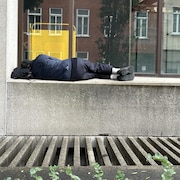 Un homme dort sur un muret de béton près d'un immeuble.