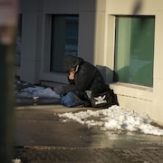 Un homme assis par terre devant un goblet en carton. Il y a de la neige autour.