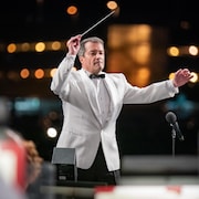 Jacques Lacombe, vêtu d'un veston blanc, tient une baguette de chef d'orchestre. 