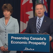 Jason Kenney parle devant un podium. La ministre se tient debout à sa gauche.