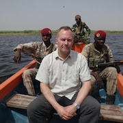 Le journaliste Jean-François Bélanger, assis dans une barque avec des militaires armés. 