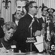 Jean-Jacques Bertrand, debout, en conférence de presse, devant de nombreux micros. Sa femme est assise à ses côtés et signe un document.