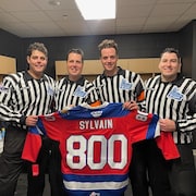 Quatre hommes habillés en arbitre de hockey tenant un gilet rouge avec le nom «Sylvain» et le chiffre «800» d'inscrit. 