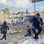 Un homme assis sur une chaise et fume, avec deux enfants à proximité, devant un immeuble détruit. 