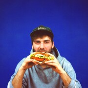 On voit le chanteur en train de manger un sandwich aux tomates, devant un fond bleu.