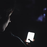 Un adolescent dans le noir, de dos, le visage penché sur un téléphone cellulaire allumé.