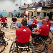 Une équipe d'athlètes de basketball en fauteuil roulant dans un gymnase écoutent leur entraîneur.