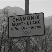 Enseigne avec l'inscription : Chamonix Mont-Blanc ville olympique. On voit des montagnes enneigées en arrière-plan.