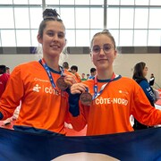 Deux jeunes filles tiennent un drapeau de la délégation de la Côte-Nord et montrent leur médaille de bronze à la caméra. 