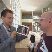 Un jeune homme a dans ses mains une photo de Roy Dupuis, qu'il présente à un autre homme. 