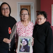 Joan Wylde, Émilie Ruperthouse-Wylde et Kathy Ruperthouse debout dans une maison. Celle du milieu tient une photographie dans ses mains.