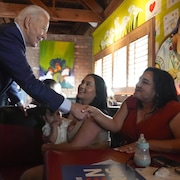Dans un restaurant, Joe Biden serre la main de deux jeunes femmes dont l'une tient dans ses bras un jeune enfant.