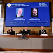 Un homme, au centre d'une femme et d'un homme, annonce les lauréats du prix Nobel de physique.