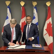 Le maire de Toronto, John Tory, écrit sur un document aux côtés du consul général devant les drapeaux du Canada et de la Ville de Toronto.
