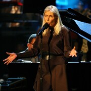 Joni Mitchell chante, les bras ouverts, accompagnée au piano par un homme.