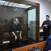 Une femme menottée dans une cage de verre; un gardien de sécurité et un avocat sont à l'extérieur. 