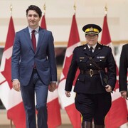 Justin Trudeau en complet et Brenda Lucki en uniforme de la GRC marchent côte à côte en direction de la caméra. Des drapeaux du Canada sont derrière eux au fond de la pièce.