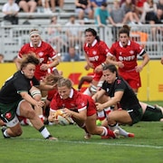 Une joueuse de rugby tient le ballon en tombant, au milieu des joueuses sur un terrain en gazon pendant un match.