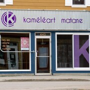 Extérieur du bureau. La lettre K est inscrite en grosse lettre dans une vitrine. Sur un panneau publicitaire au-dessus de la porte d'entrée, il est écrit : Kaméléart Matane.