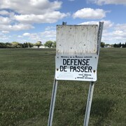 Un panneau qui dit "défense de passer, ministère de la Défense nationale", planté sur l'herbe dans un grand terrain vague.