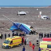 Des camions et un hélicoptère immobilisé sont à proximité de l'épave d'un avion écrasé. Des gens discutent parmi les décombres.