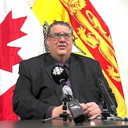 Kelly Lamrock assis à une table, un micro devant lui, devant un mur blanc où sont agrafés de travers des drapeaux du Canada et du Nouveau-Brunswick.