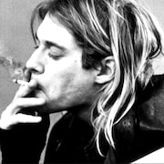 Kurt Cobain fume une cigarette appuyé sur sa guitare. 