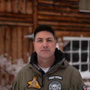 Portrait de Patrick Lemay à l'extérieur de sa cabane au Yukon. Il porte un manteau rempli d'écussons.